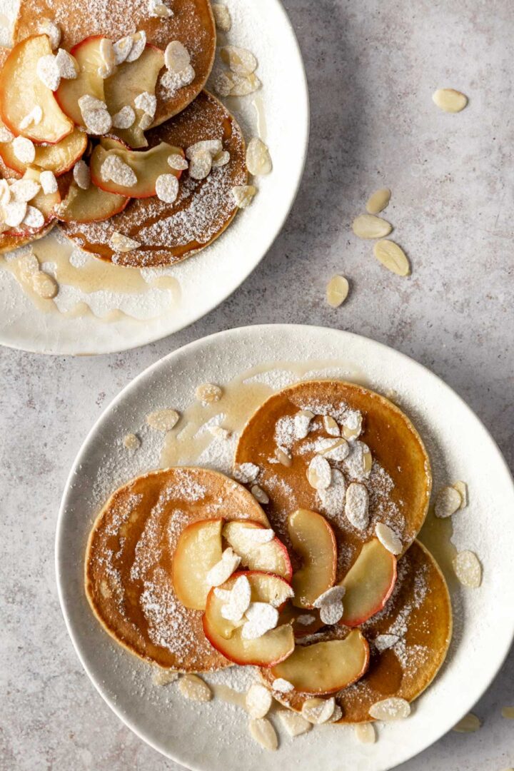 two plates with 3 pancakes each, baked apple slices, almond flakes, maple syrup and icing sugar on top