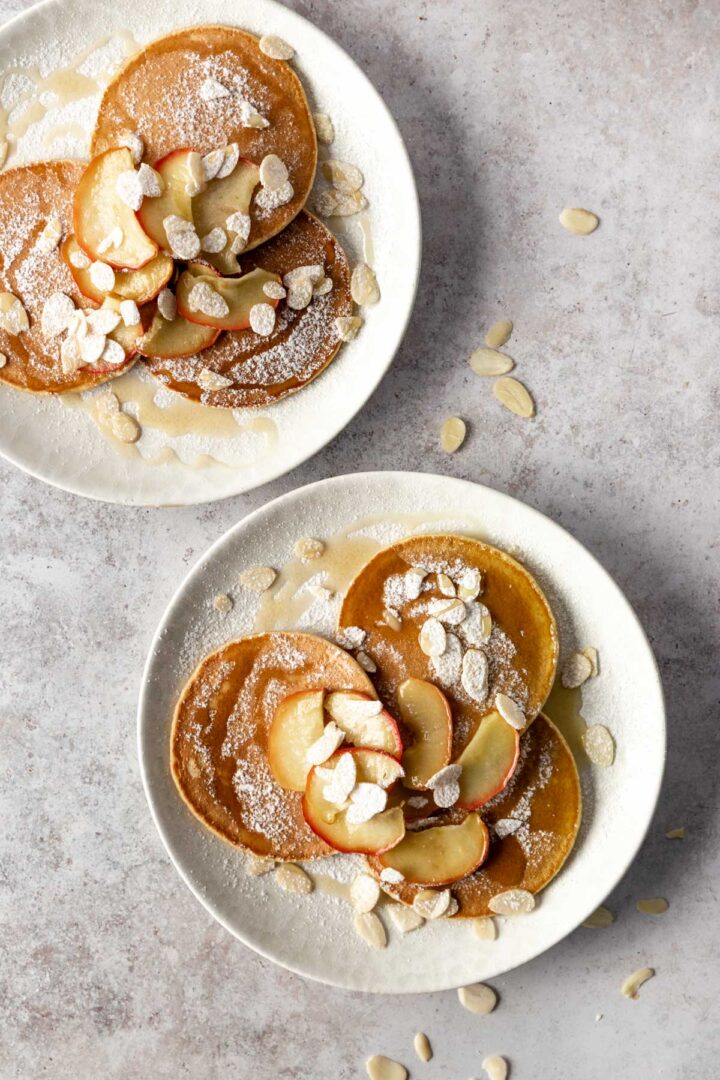two plates with 3 pancakes each, baked apple slices, laminated almonds, maple syrup and icing sugar on top