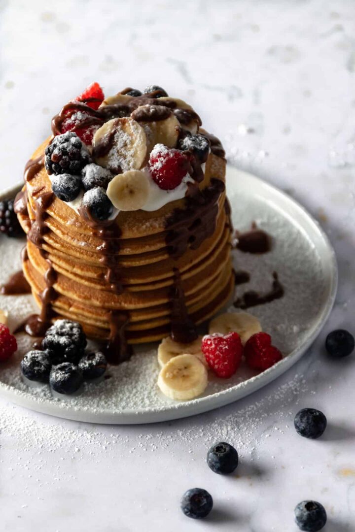 a plate with a stack of pancakes, coconut yogurt, sliced banana, berries, chocolate ganache and powdered sugar on top