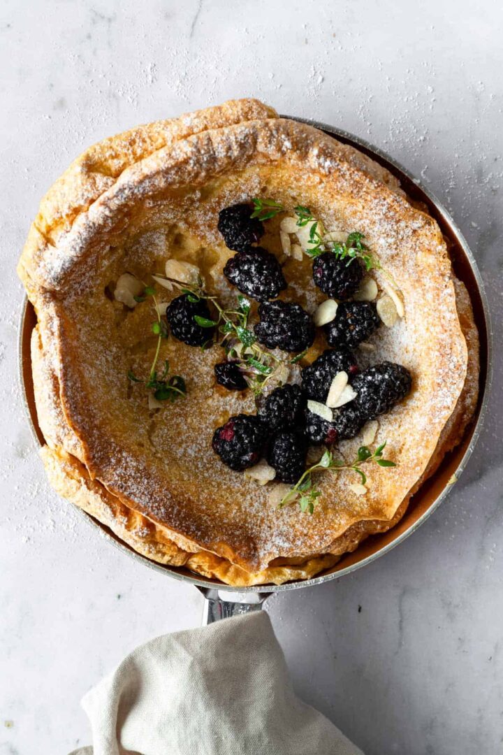dutch baby pancake on a skillet with blackberries, thyme, powdered sugar and almond flakes on top