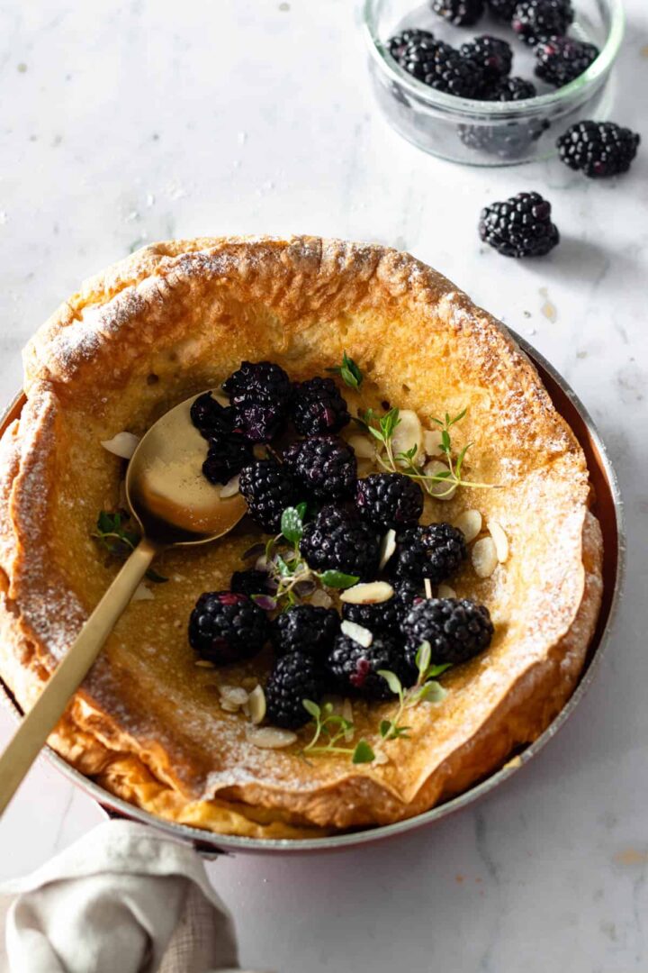 pancake on a skillet with a spoon on top and a small glass bowl with berries on the side