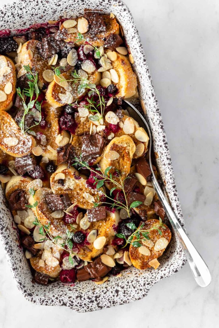 a casserole with sliced bread, chocolate chunks, flaked almonds, berries, icing sugar and thyme on top with a silver serving spoon on the side