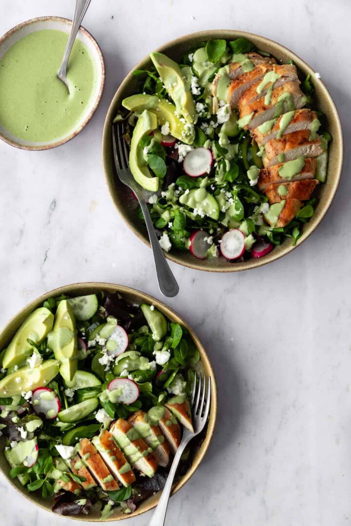 two bowls with sliced chicken breast, sliced avocado, radishes, feta cheese, cucumber, salad mixture, green dressing on top and a small bowl with green cream dressing on the side