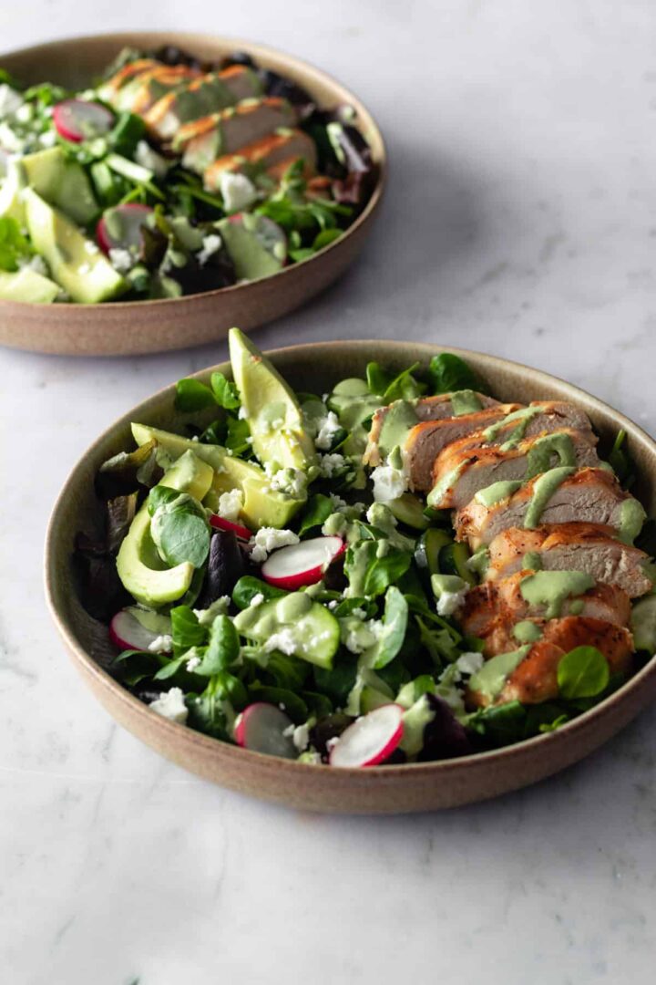 two bowls with sliced chicken breast, sliced avocado, radishes, feta cheese, cucumber, salad mixture and green dressing on top