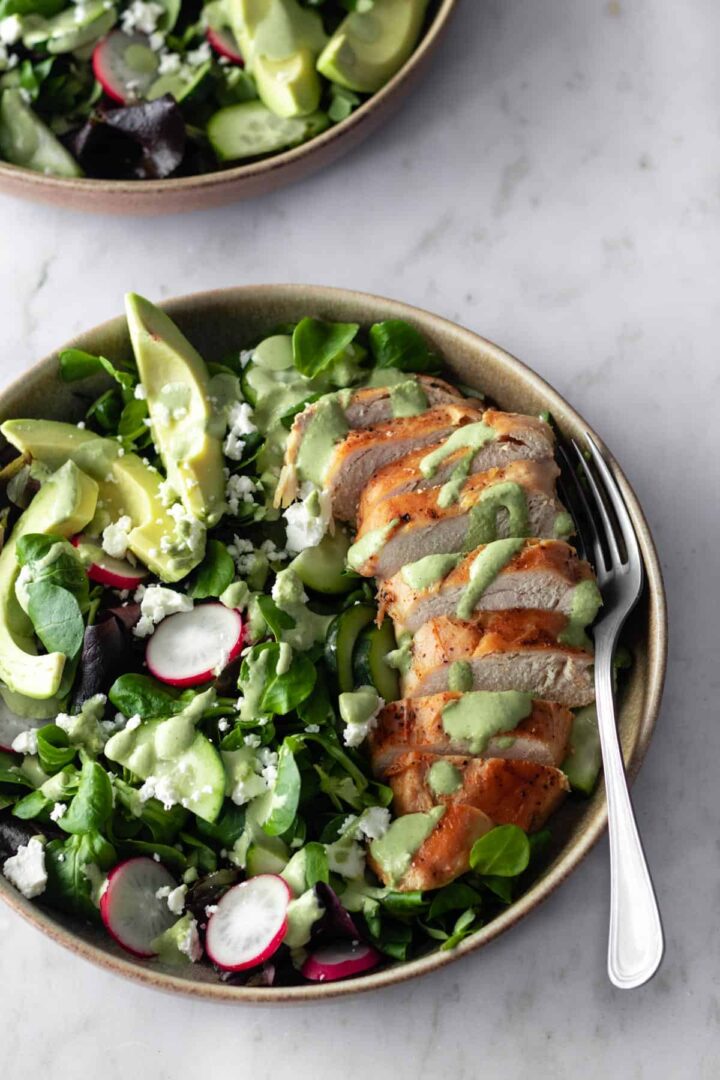 a bowl with sliced chicken breast, sliced avocado, radishes, feta cheese, cucumber, salad mixture and green dressing on top
