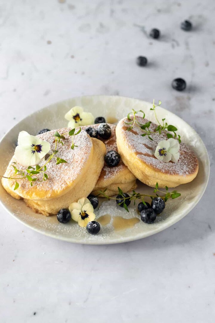 a plate with three Japanese pancakes, blueberries, thyme, edible flowers, maple syrup and powdered sugar on top and blueberries on a marble table