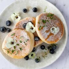 a plate with three pancakes, blueberries, thyme, edible flowers, maple syrup and powdered sugar on top
