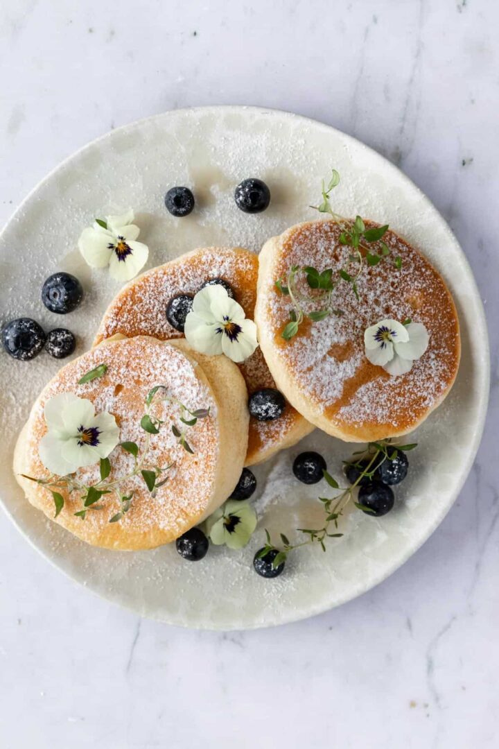 a plate with three pancakes, blueberries, thyme, edible flowers, maple syrup and powdered sugar on top