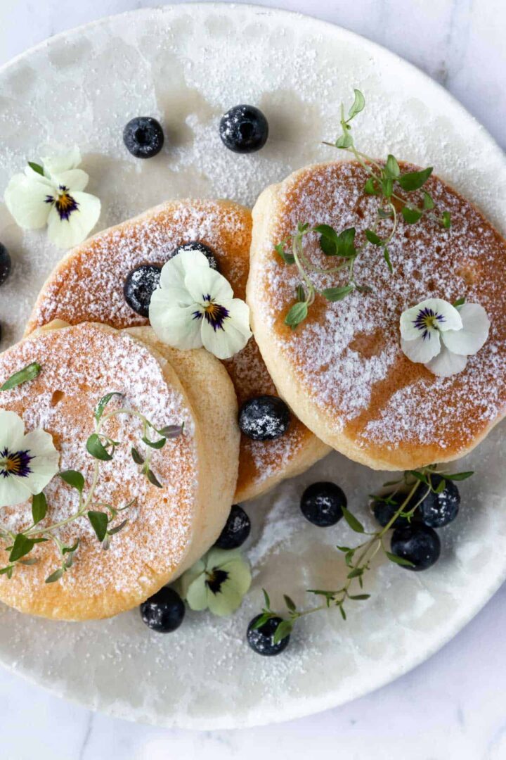a plate with three Japanese pancakes, blueberries, thyme, edible flowers, maple syrup and powdered sugar on top