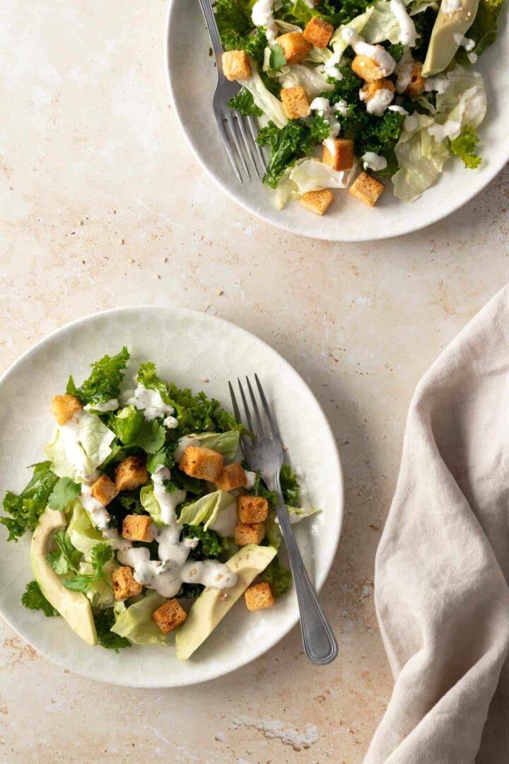two plates served with kale, iceberg lettuce, sliced avocado, bread croutons, sesame seeds, with dressing and a bege kitchen towel on the side