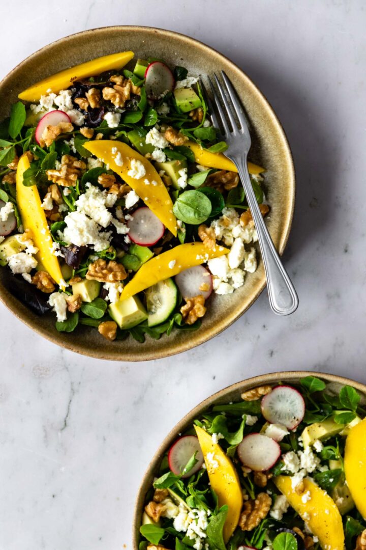 two bowls with mixed greens, sliced radishes, sliced mango, cucumber, chopped walnuts, feta cheese and avocado on top of a marble table