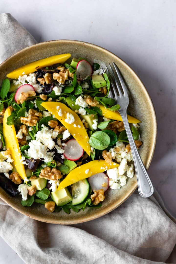 a bowl with mixed greens, cubed avocado, sliced radishes, sliced cucumber, chopped walnuts, mango and crumbled feta cheese on top of a beige kitchen towel