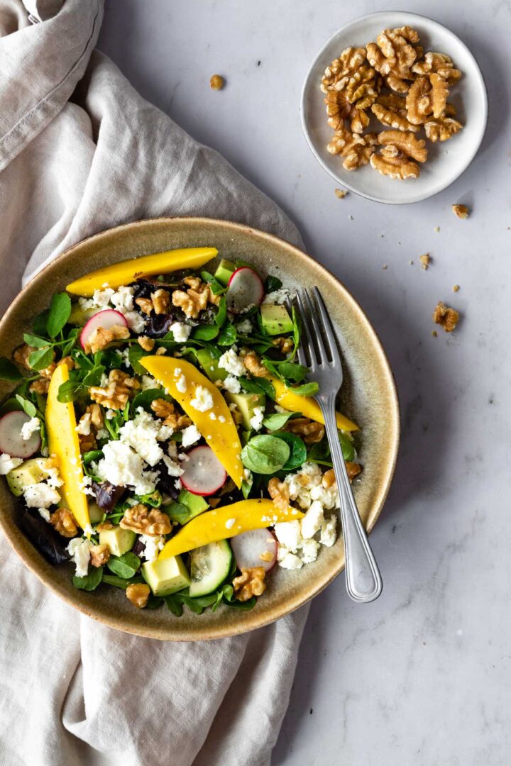 a bowl with mixed greens, cubed avocado, sliced radishes, sliced cucumber, chopped walnuts, mango and crumbled feta cheese on top of a beige kitchen towel. A small bowl with chopped walnuts on the side