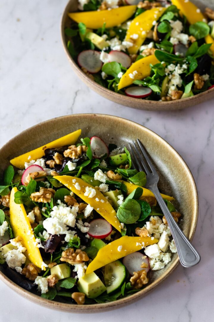 two bowls with mixed greens, sliced radishes, sliced mango, cucumber, chopped walnuts, feta cheese and avocado on top of a marble table