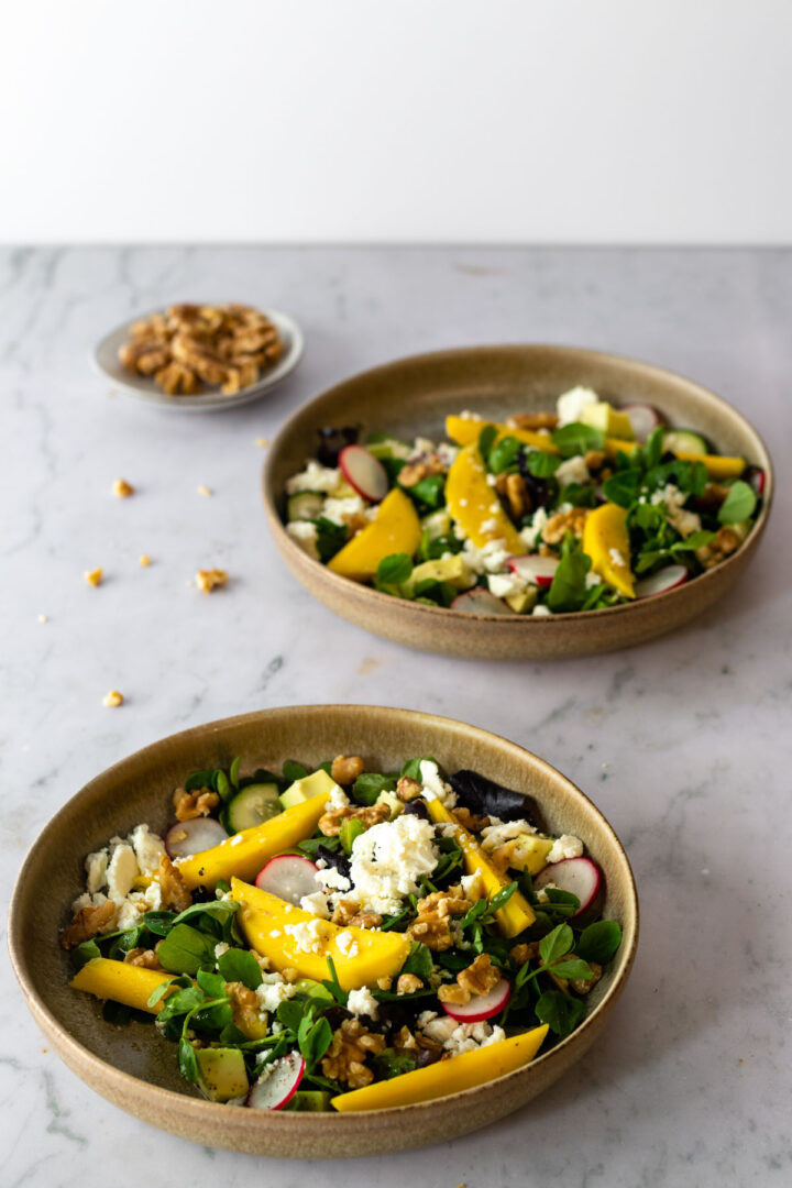 two bowls with mixed greens, sliced radishes, sliced mango, cucumber, chopped walnuts, feta cheese and avocado on top of a marble table. A small bowl with walnuts out of focus