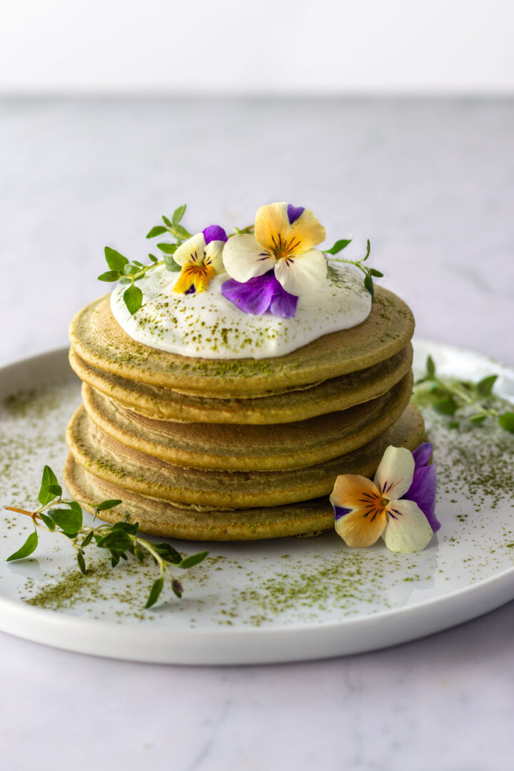 a plate with a stack of pancakes, coconut yogurt, edible flowers and match powder on top