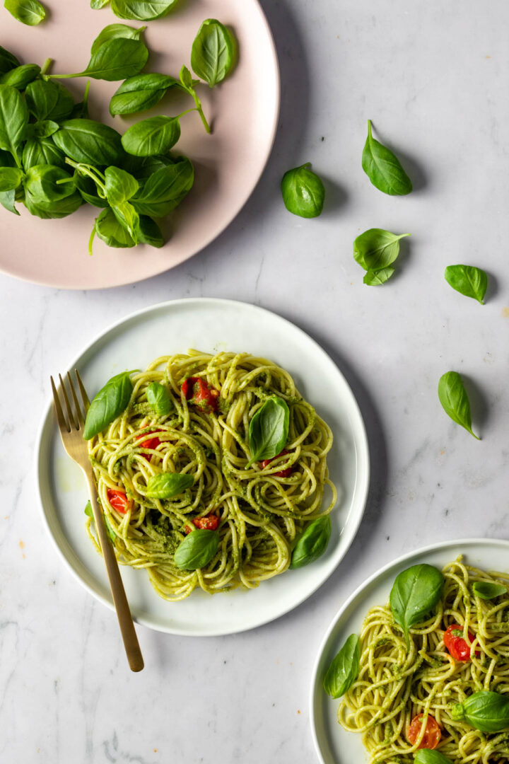 two white plates with pesto pasta, basil leaves and cherry tomatoes. A pink plate with more basil. Basil leaves on top of the marble table