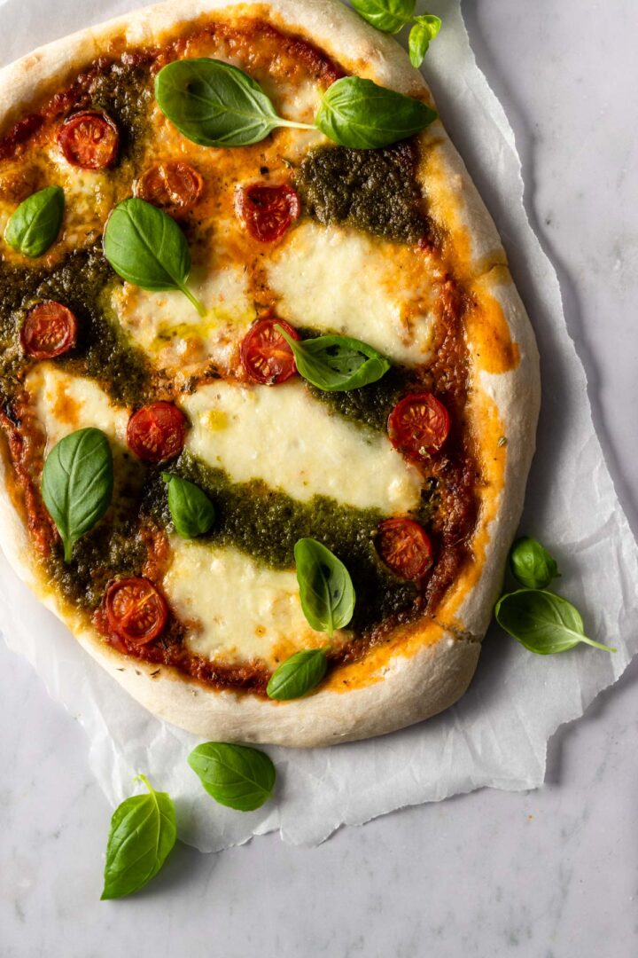 pizza on top of parchment paper with mozzarella, tomato sauce, pesto sauce, cherry tomatoes and basil leaves
