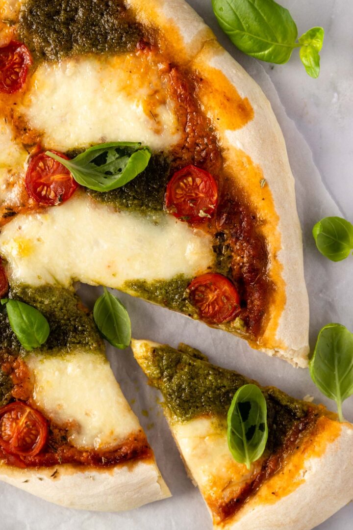 a pizza sliced with mozzarella, tomato sauce, pesto sauce, cherry tomatoes and basil leaves