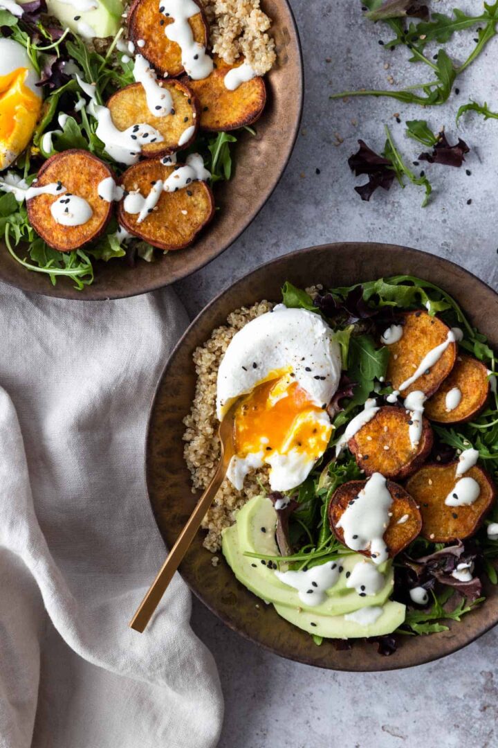 two bowls with mixed greens, quinoa, sliced avocado, sweet potato, poach egg and dressing on top