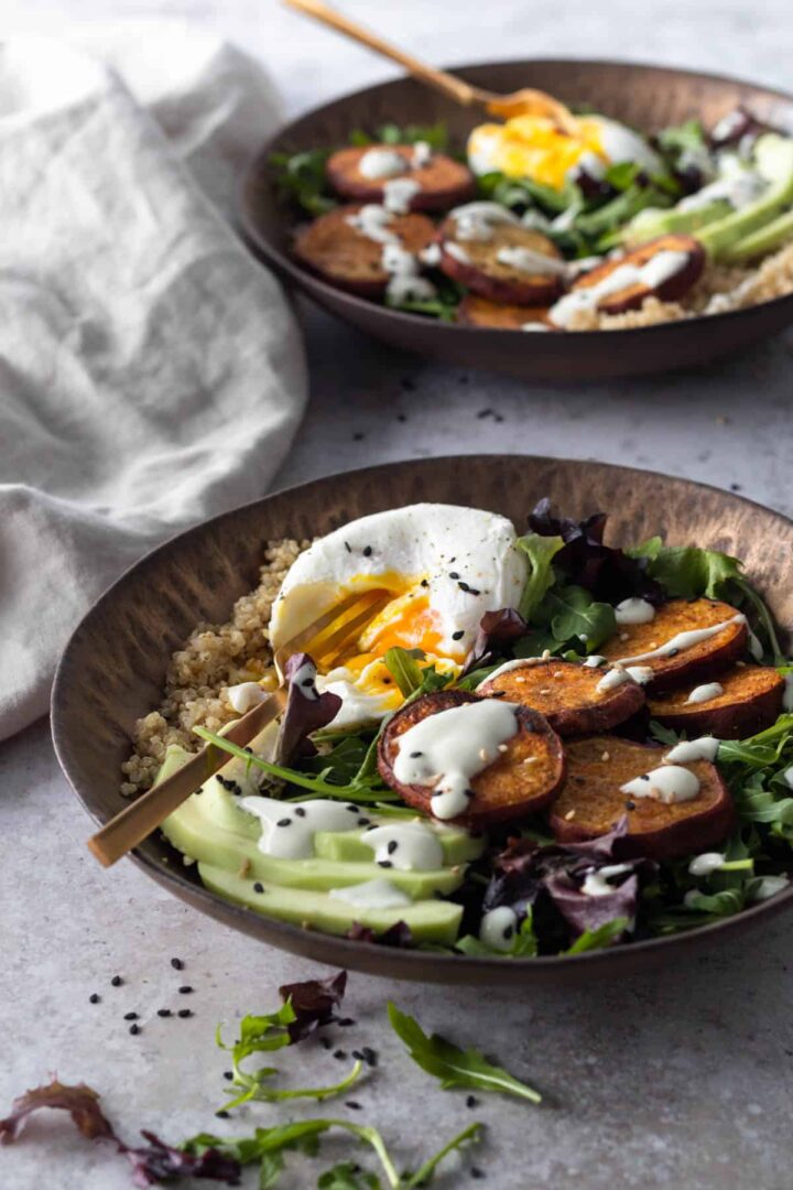 two bowls with mixed greens, quinoa, sliced avocado, sweet potato, poach egg and dressing on top