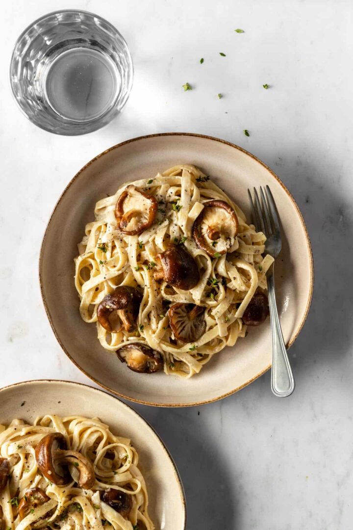 Shiitake mushrooms and fettuccine pasta served on a plate, garnished with fresh thyme and a glass with water on the side