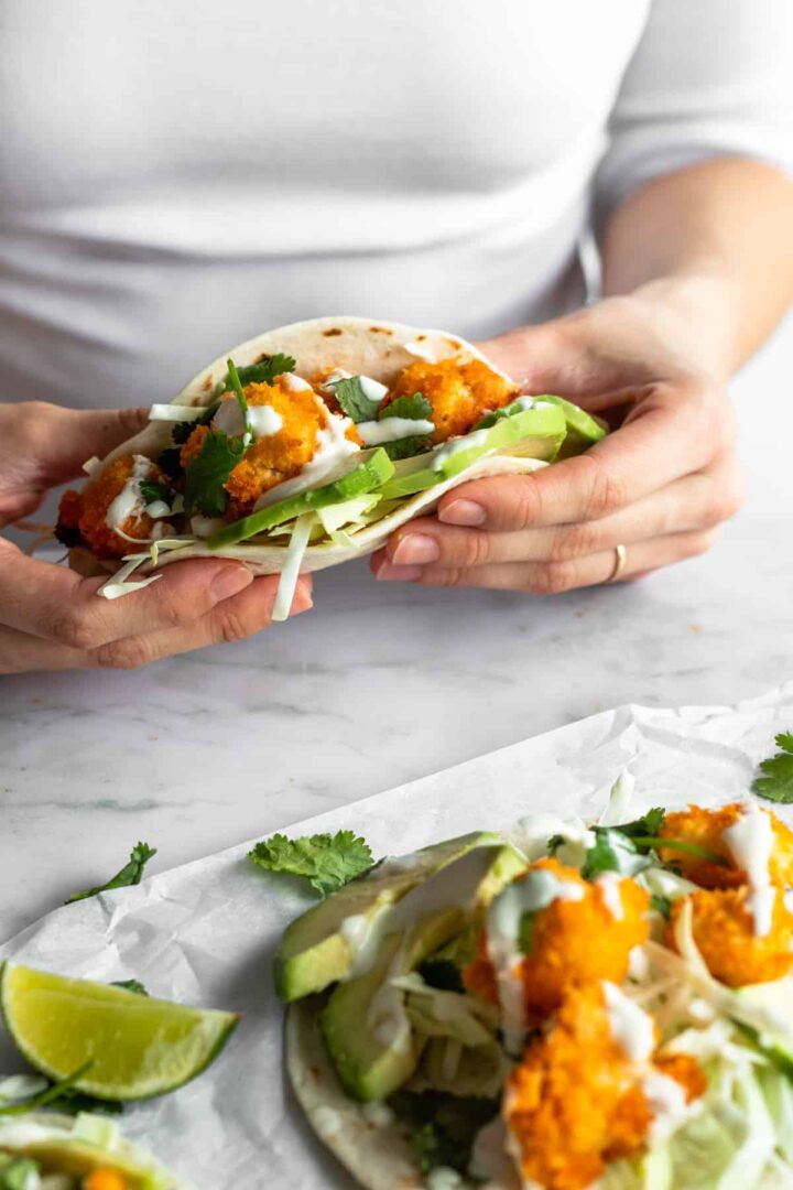 a person holding a taco with cauliflower bites, shredded cabbage, coriander, avocado and a creamy sauce. More cauliflower tacos and lime wedges on top of a marble table