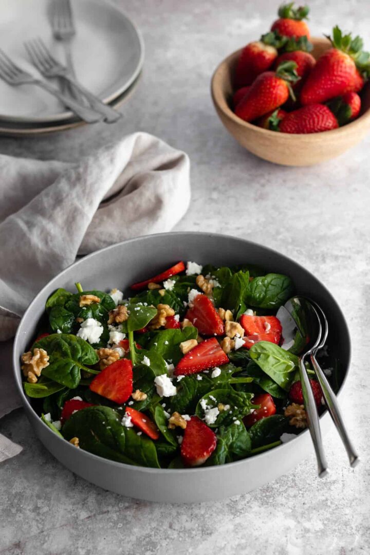 a grey bowl with spinach, strawberries, feta cheese and walnuts. A small bowl with more strawberries, two plates and three forks and a kitchen towel on top of a backdrop