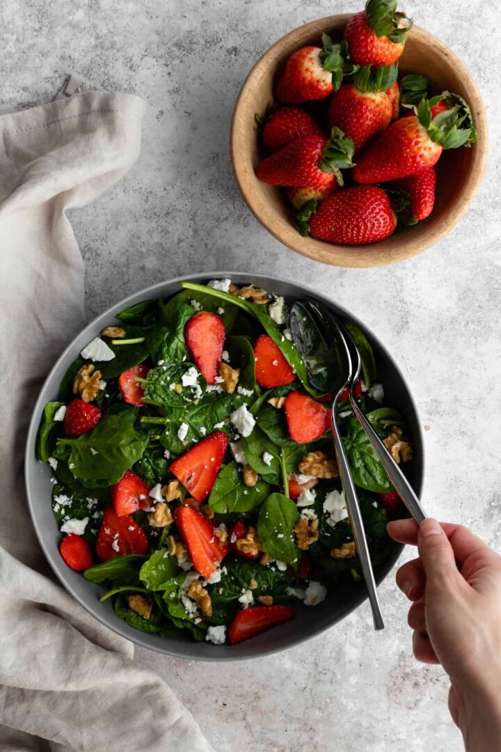 a grey bowl with spinach, strawberries, feta cheese and walnuts. A small bowl with more strawberries, two plates and three forks and a kitchen towel on top of a backdrop. One hand holding a serving fork