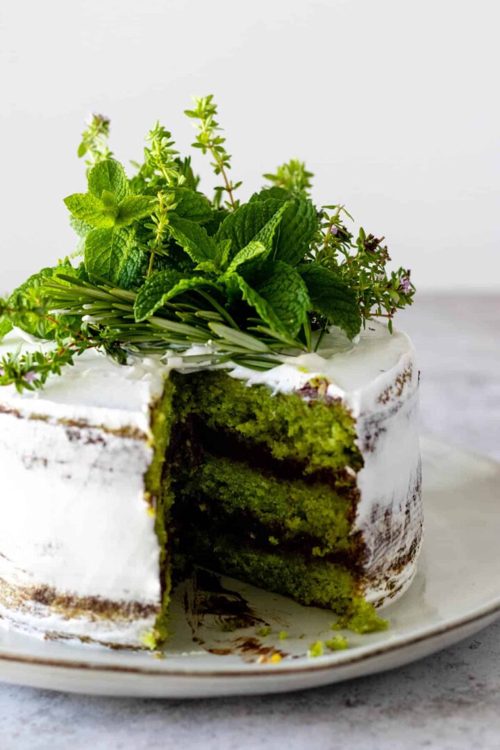 a sliced three layered green cake covered with whipping cream frosting, with a chocolate ganache and decorated with fresh herbs like mint, rosemary and thyme