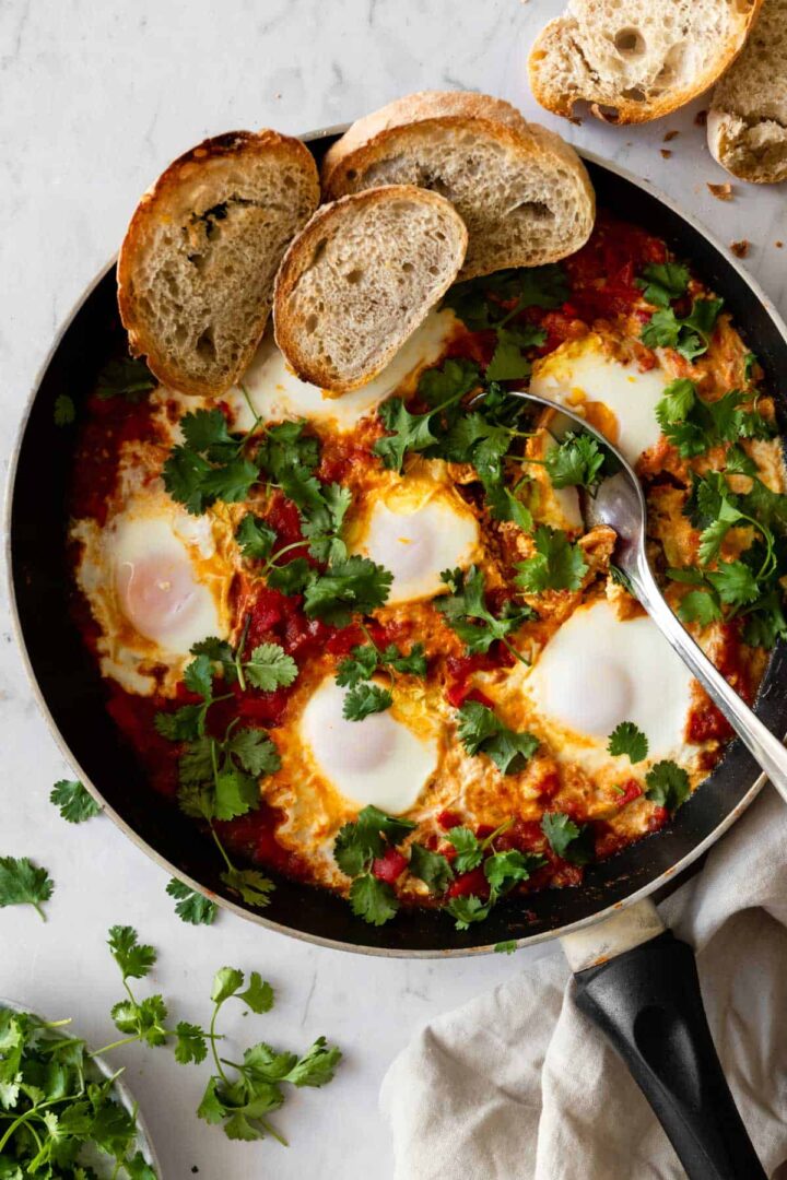 a kitchen pan with shakshuka garnished with coriander and toasted sliced bread on the side