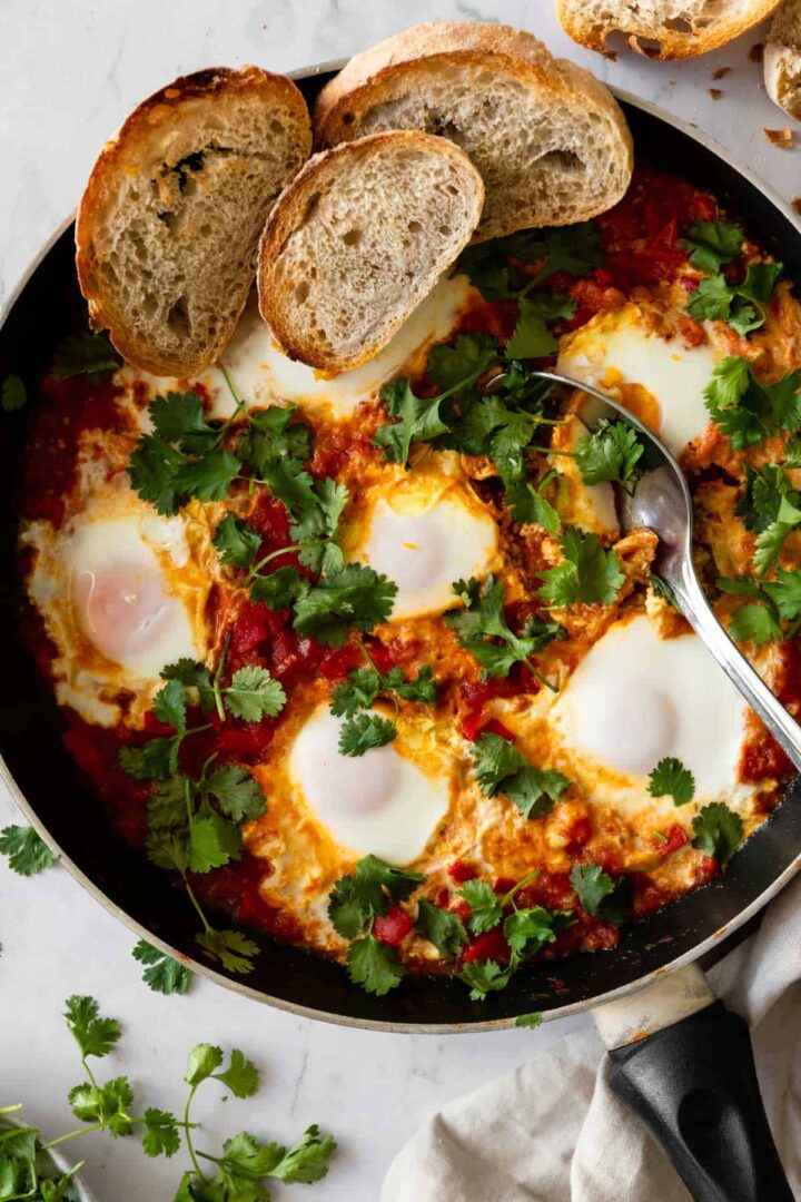a pan with shakshuka garnished with fresh coriander and sliced bread on top