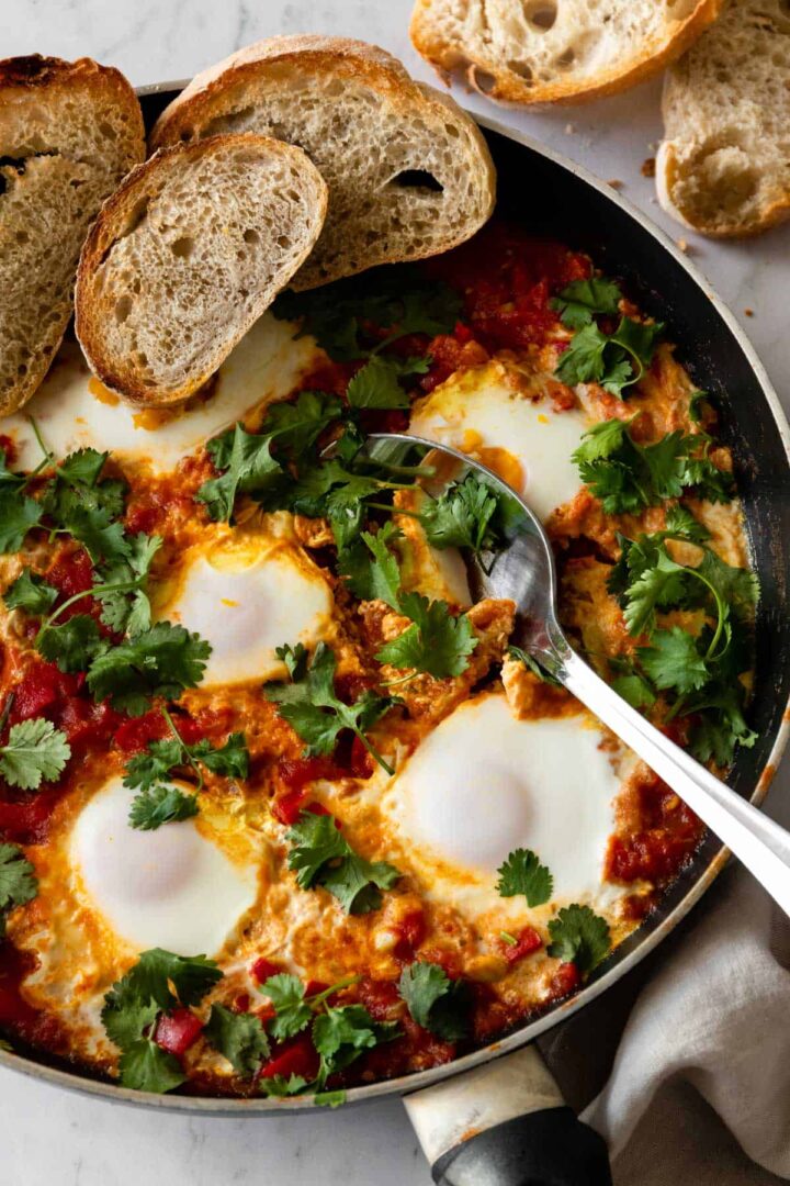 close-up of a pan with shakshuka garnished with fresh coriander and sliced bread