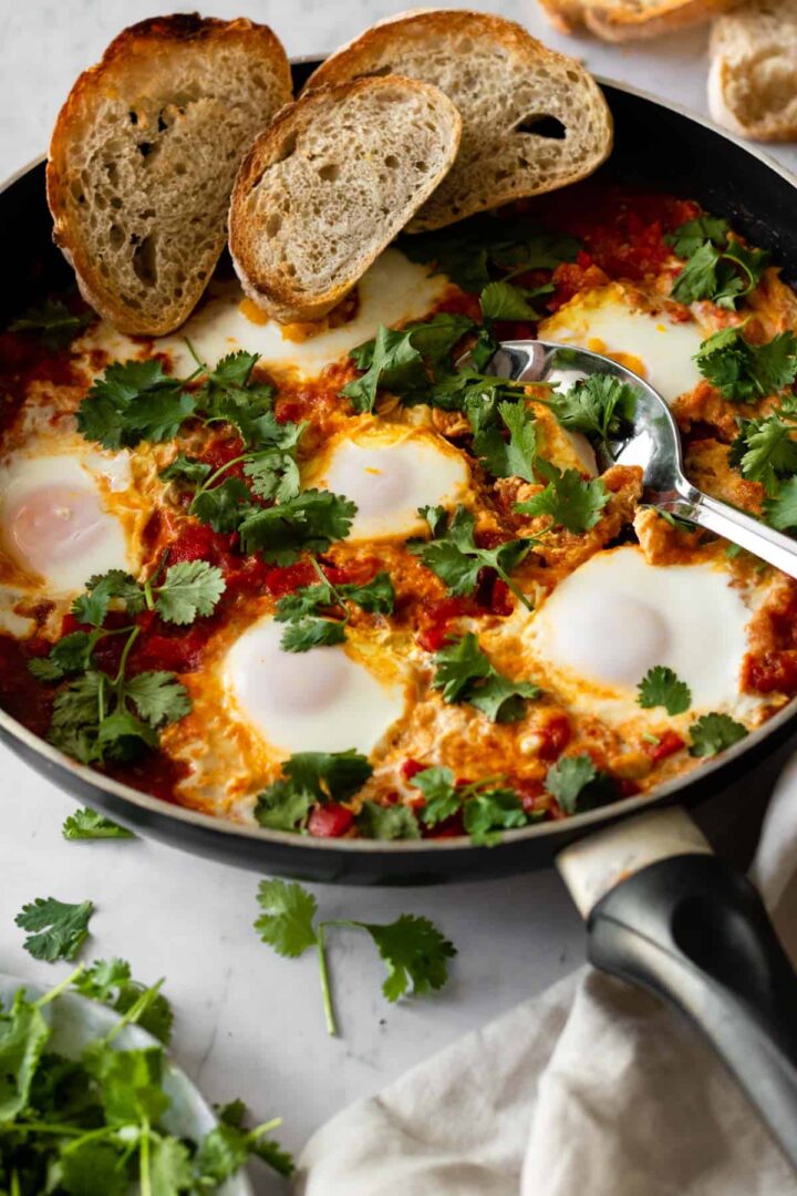 a pan with shakshuka garnished with fresh coriander and sliced bread on top