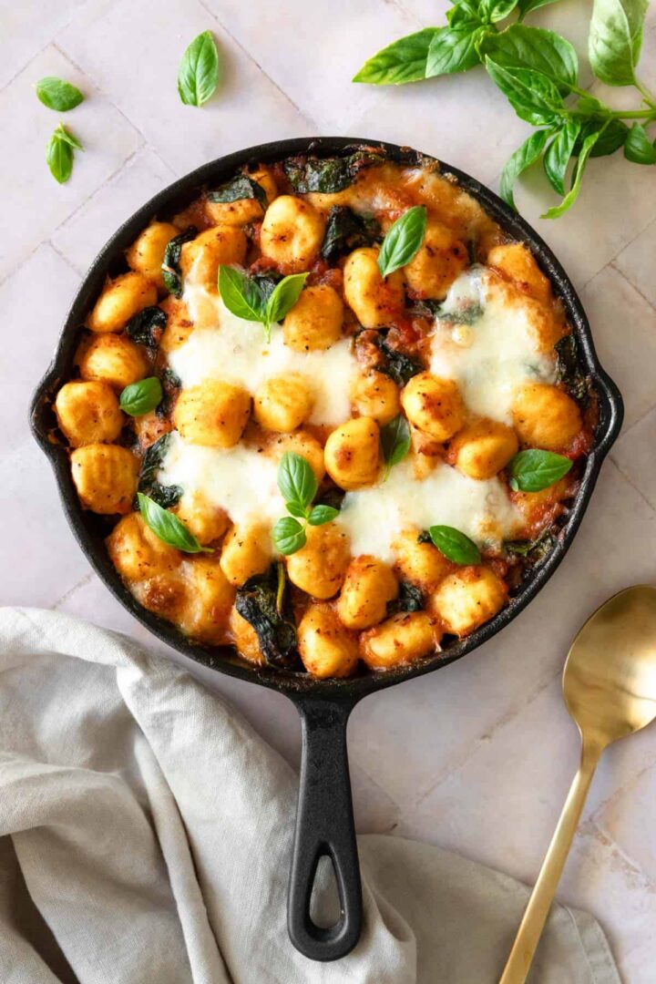 a skillet with gnocchi bake, garnished with fresh basil and a gold spoon on the side