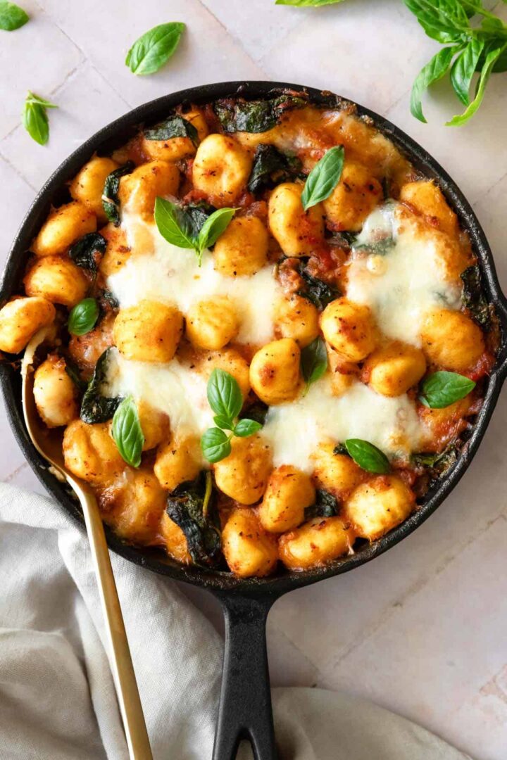 a skillet with gnocchi bake, garnished with fresh basil and a gold spoon on the side