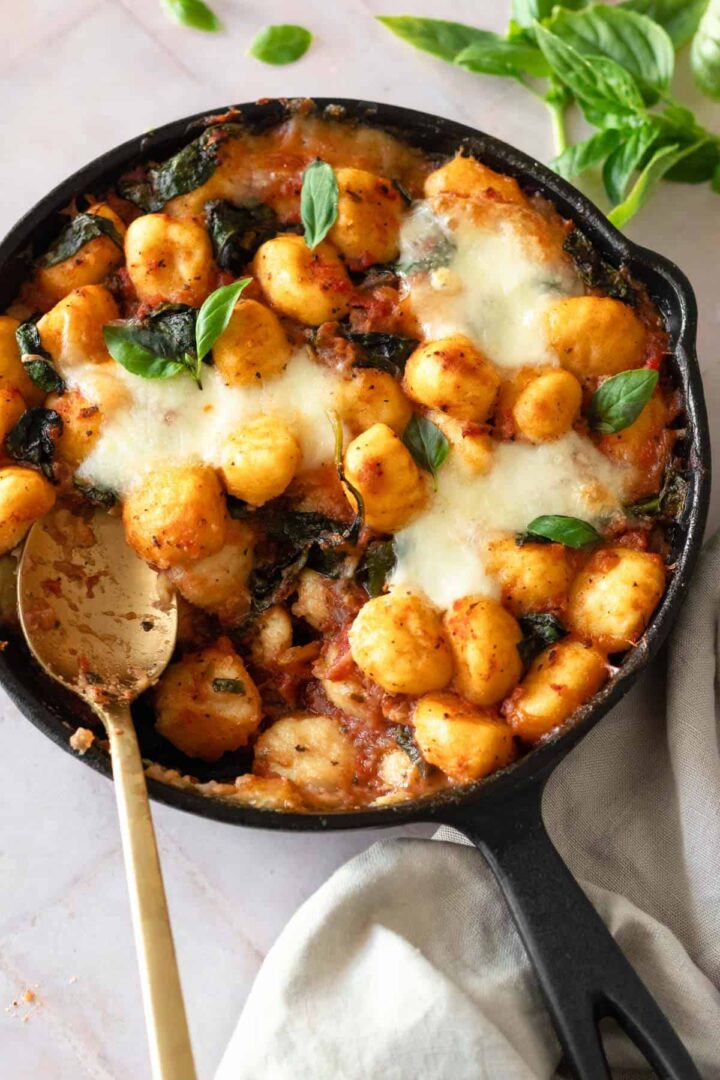 a skillet with gnocchi bake, garnished with fresh basil and a gold spoon on the side