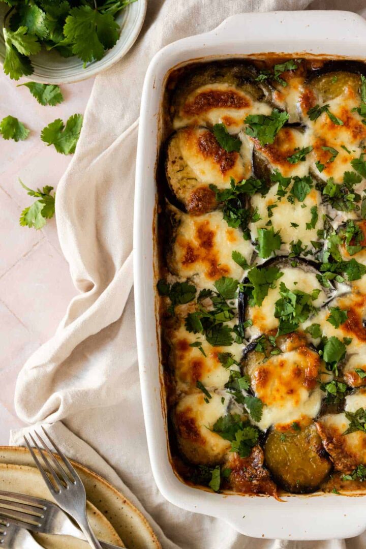 vegetable moussaka on a baking dish with chopped coriander on top, a bowl with more coriander on the side and forks on the bottom left corner