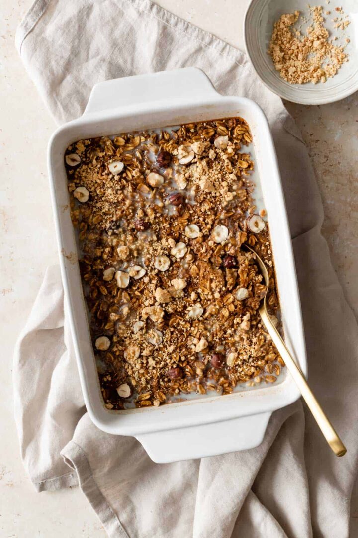 pumpkin baked oatmeal on a baking dish with a gold spoon on the side. A small bowl with paΓ§oca on the top right corner