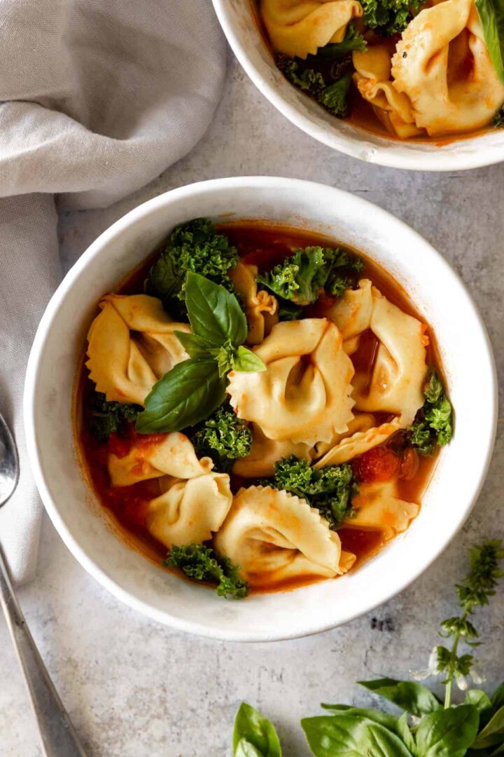 a bowl served with tortellini soup with kale, tomatoes and garnished with fresh basil. Another bowl on the top right corner with the same soup.