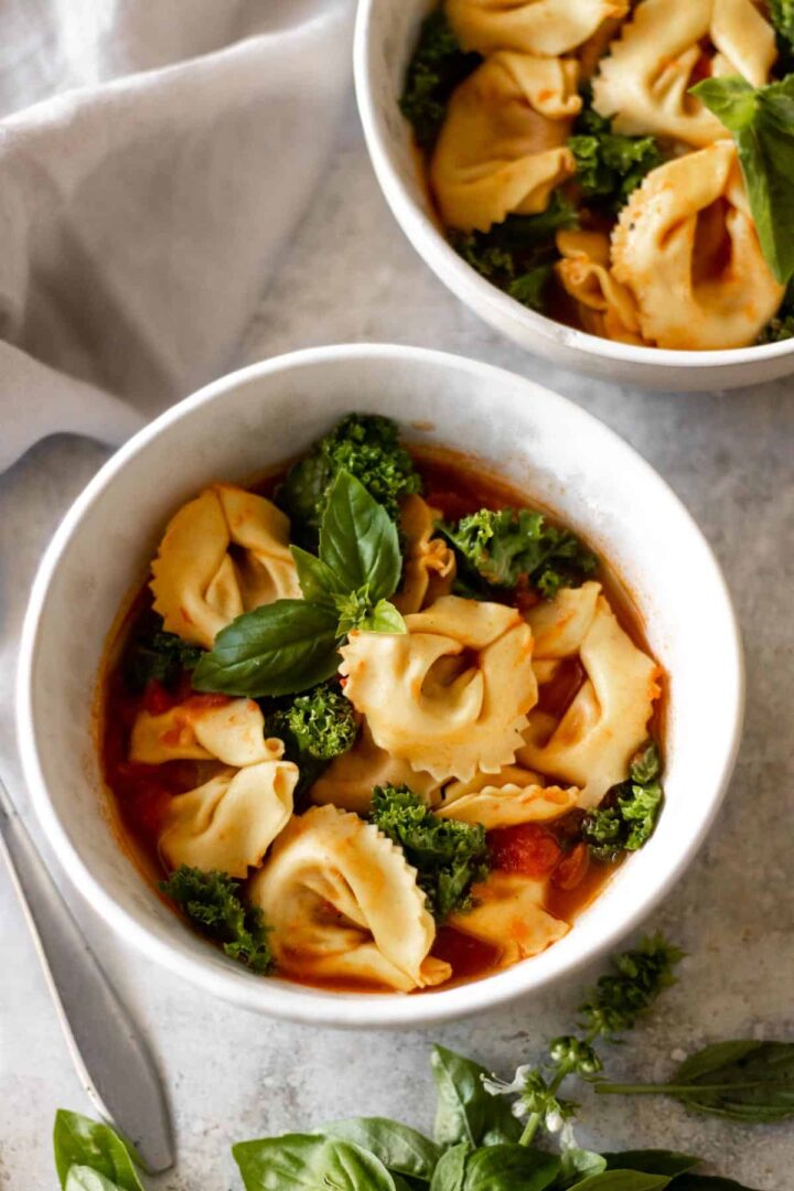 two bowls served with tortellini soup with kale, tomatoes and garnished with fresh basil