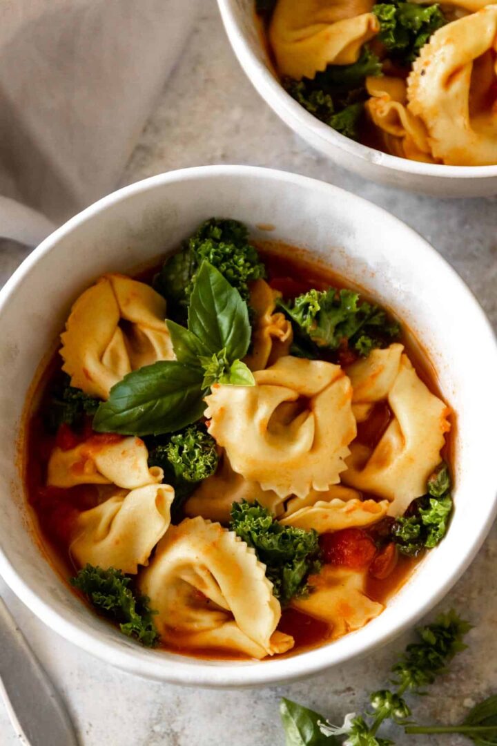 two bowls served with tortellini soup with kale, tomatoes and garnished with fresh basil