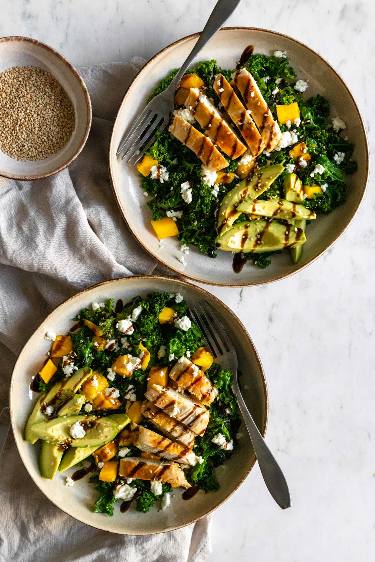 two plates with kale, chicken, avocado, feta cheese, mango, sesame seeds, and a bowl with sesame seeds on the side