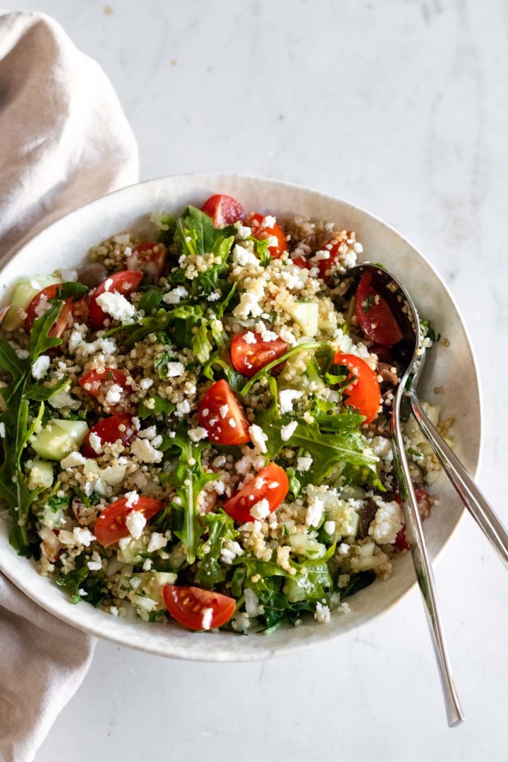 a bowl with quinoa salad on top of a marble table, two serving spoons and a kitchen towel