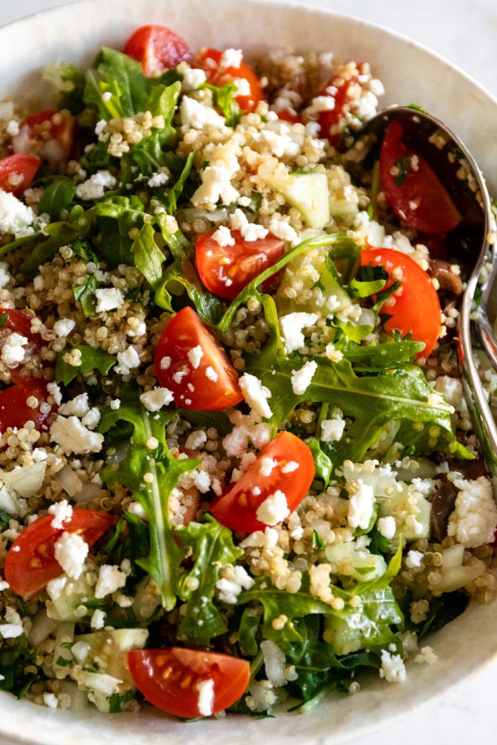 close-up of a bowl with quinoa, cherry tomatoes, arugula, crumbled feta cheese, cucumber, onion, black olives and parsley