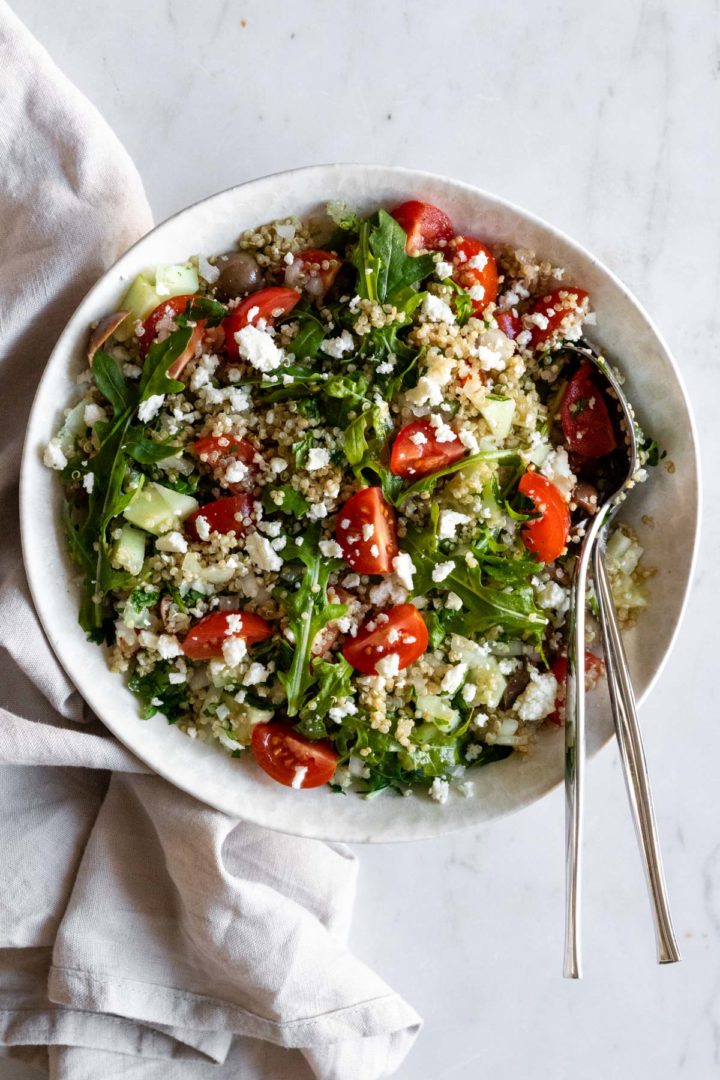 a bowl with quinoa salad on top of a marble table, two serving spoons and a kitchen towel