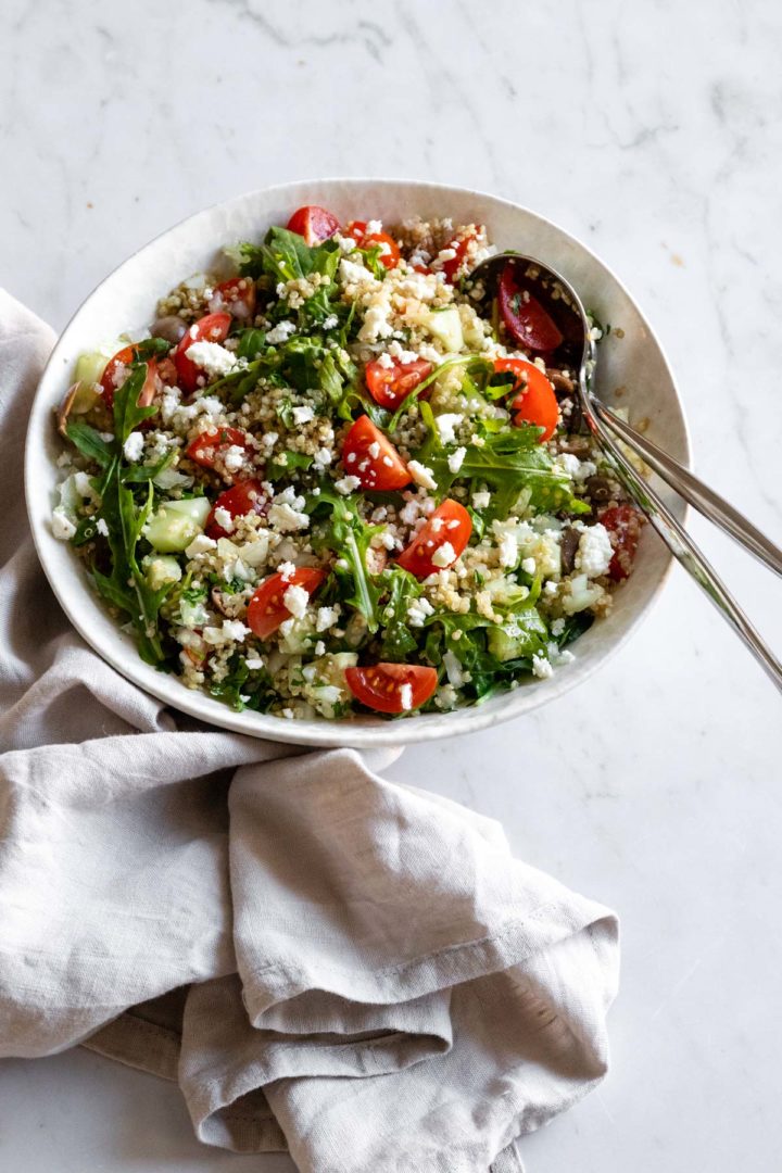 a bowl with quinoa salad on top of a marble table, two serving spoons and a kitchen towel