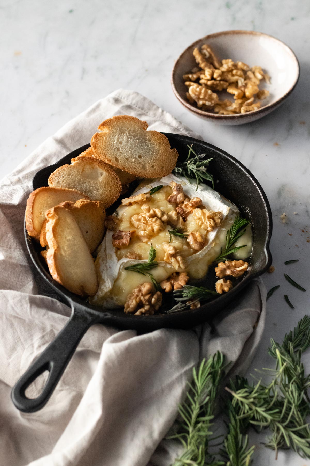 baked brie, toasted bread, walnuts and rosemary inside a black skillet, more rosemary and walnuts on the side
