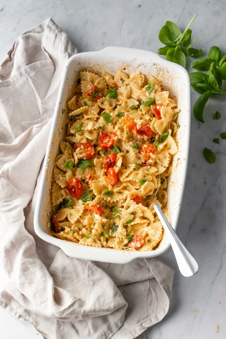 a baking dish with pasta, cherry tomatoes, chopped coriander and a spoon. More fresh basil leaves and a kitchen towel on top of a marble table