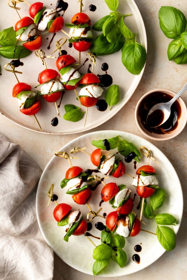 two plates served with caprese skewers garnished with blasamic glazed and basil leaves. A small bowl with a spoon and more balsamic glaze on the side